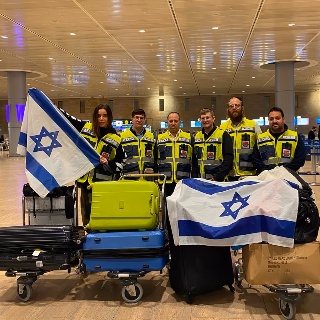 Group of six rescue personnel in yellow and black ZAKA vests standing behind luggage carts with Israeli flags at an airport.