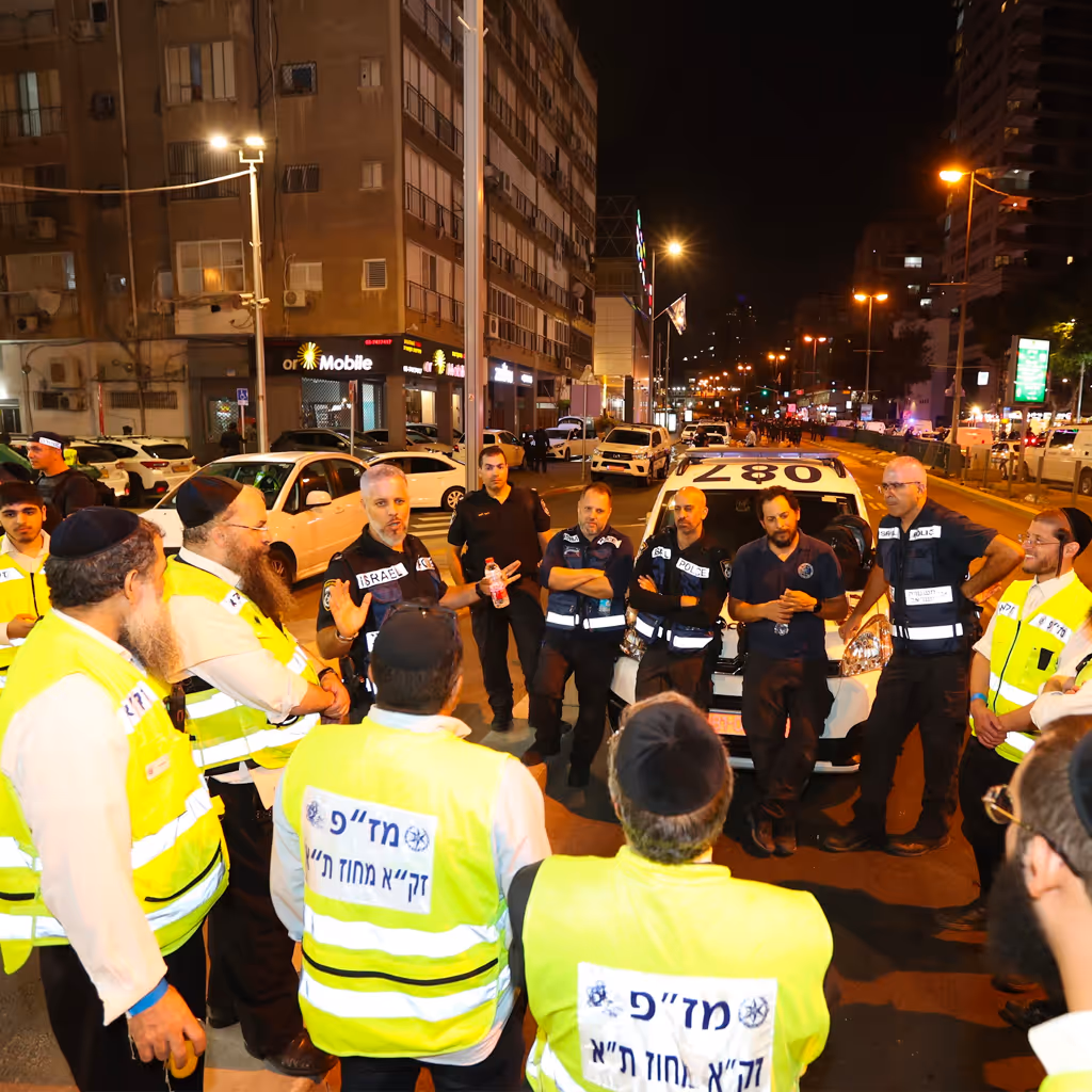 Group of emergency responders and police officers in reflective vests standing in a circle at night on a city street near a police vehicle.