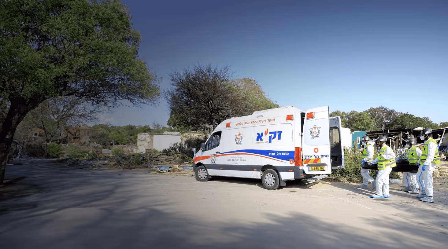 Emergency response team wearing protective suits and masks carrying a stretcher toward a ZAKA Tel Aviv ambulance on a sunny day.
