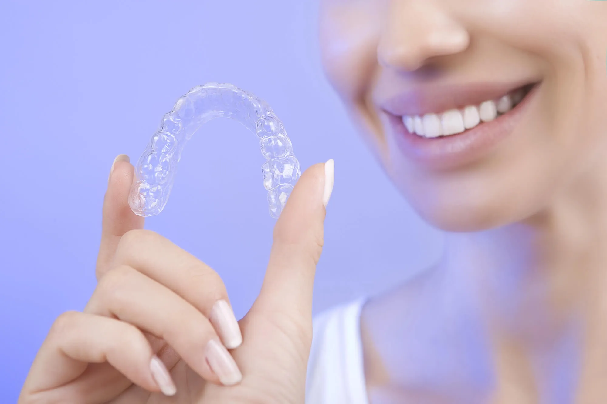 Clear aligner held between fingers in front of a smiling patient, representing removable orthodontic treatment for teeth straightening.