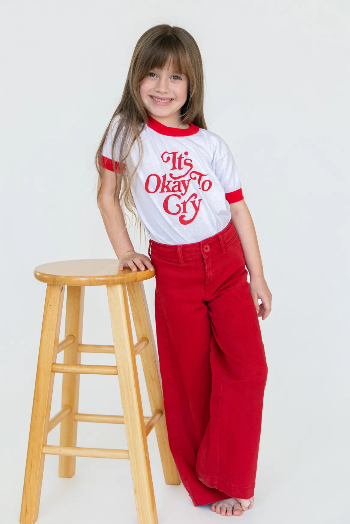 Smiling young girl with long brown hair wearing a white and red shirt that says 'It's Okay To Cry' and red pants, standing barefoot beside a wooden stool.