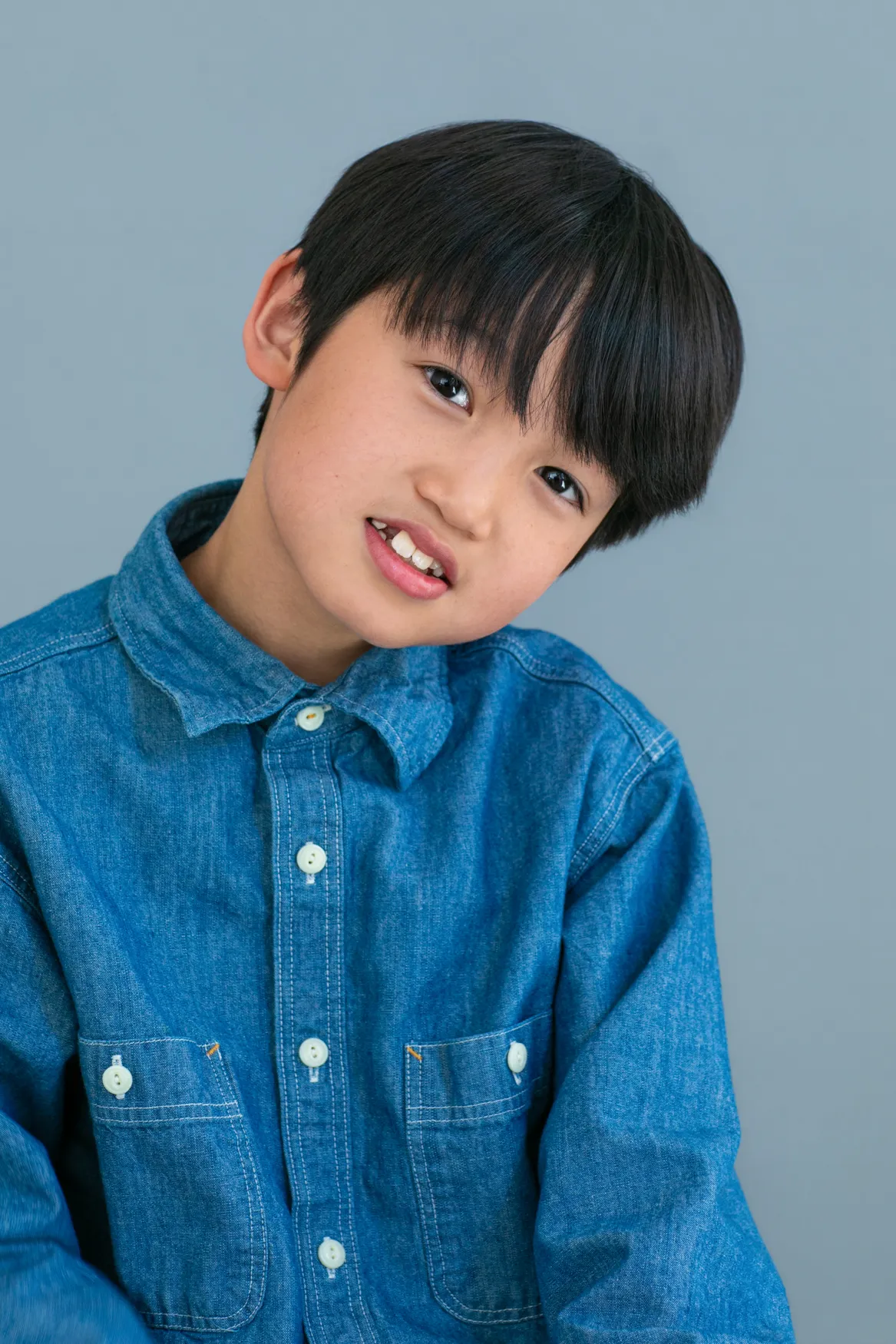 Young boy with black hair wearing a blue denim shirt against a plain grey background.