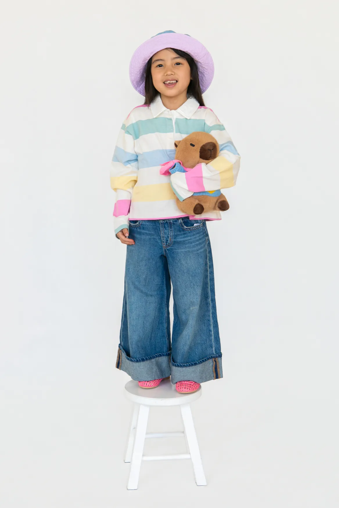 Smiling child standing on a white stool wearing a pastel-striped sweater, wide jeans, purple hat, and holding a brown stuffed animal.