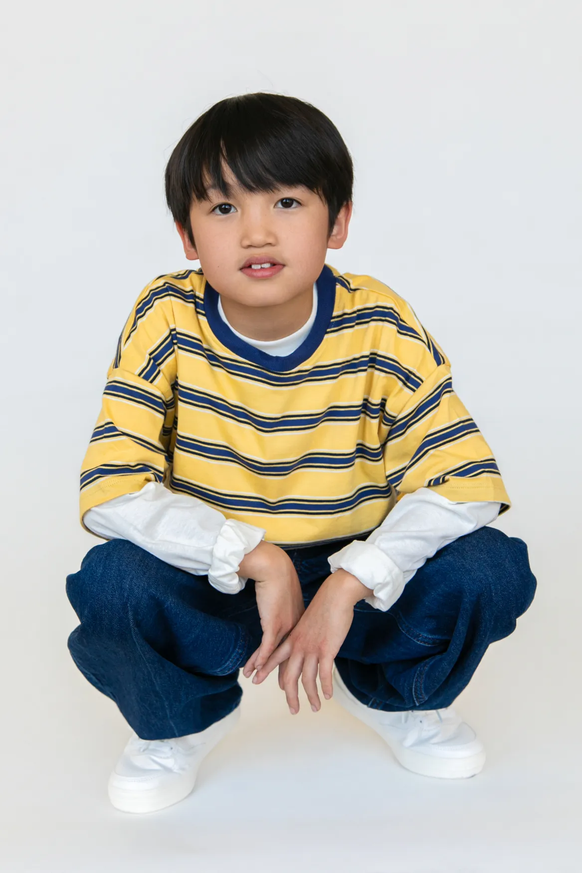 Young boy with black hair wearing a yellow and blue striped shirt, white long sleeves, blue jeans, and white sneakers squatting.
