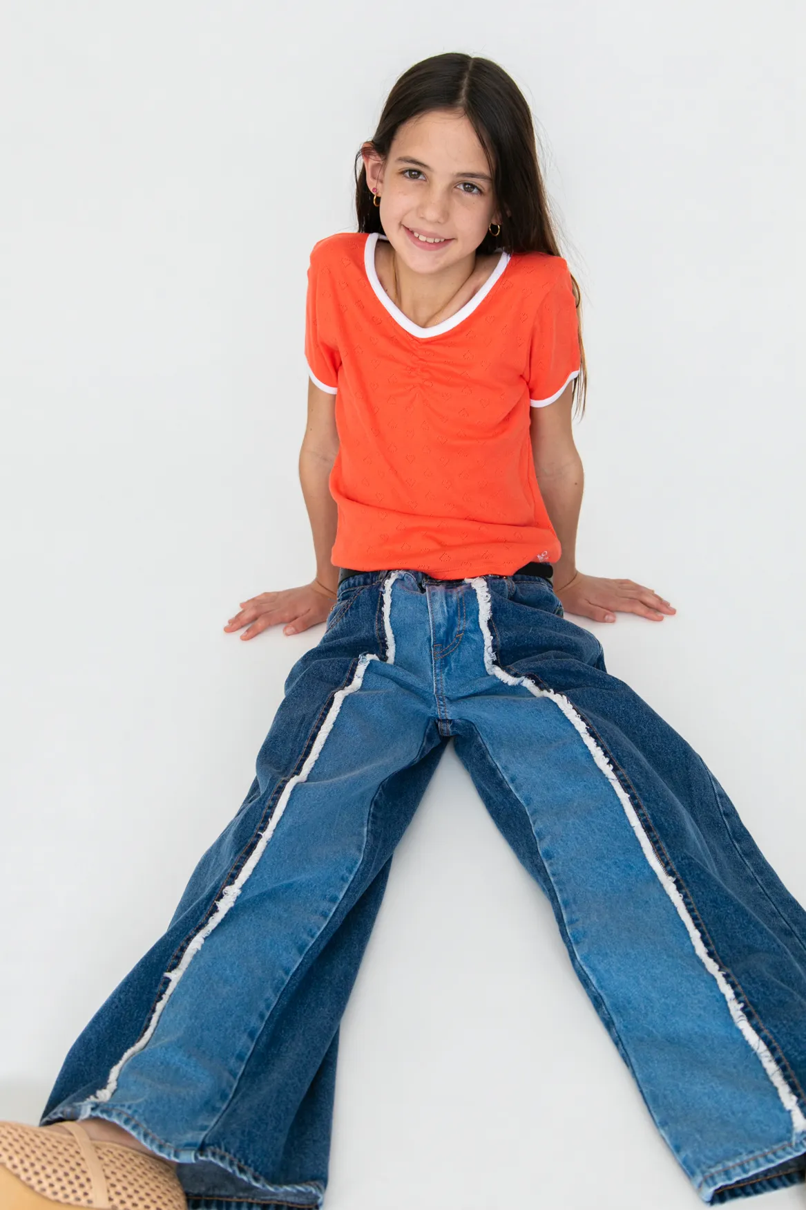 Smiling girl with long dark hair wearing an orange short-sleeve shirt and two-tone blue jeans sitting on a white floor.