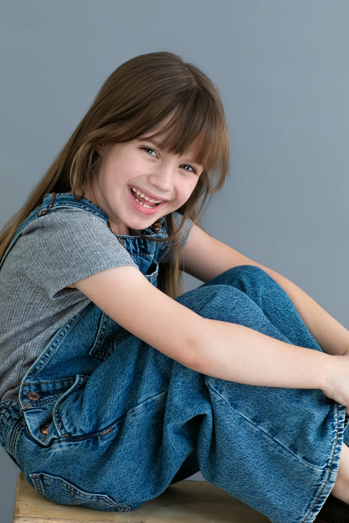 Smiling young girl with long brown hair wearing denim overalls and a gray shirt sitting with knees folded on a wooden surface.
