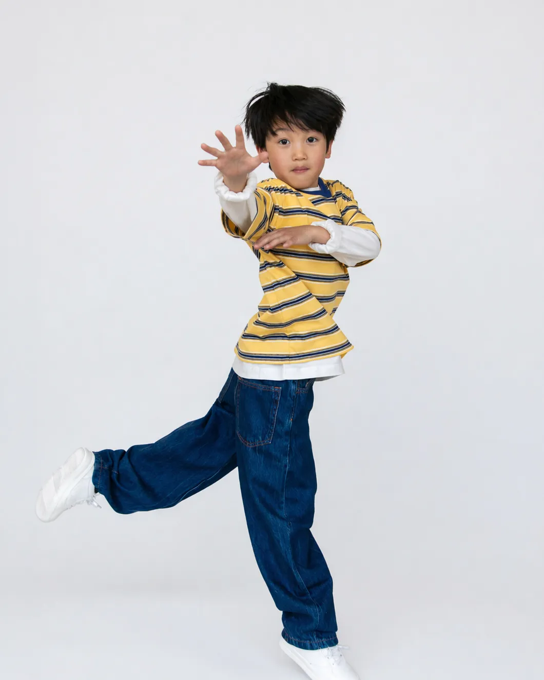 Child in yellow striped shirt and blue jeans balancing on one leg with one arm extended forward.