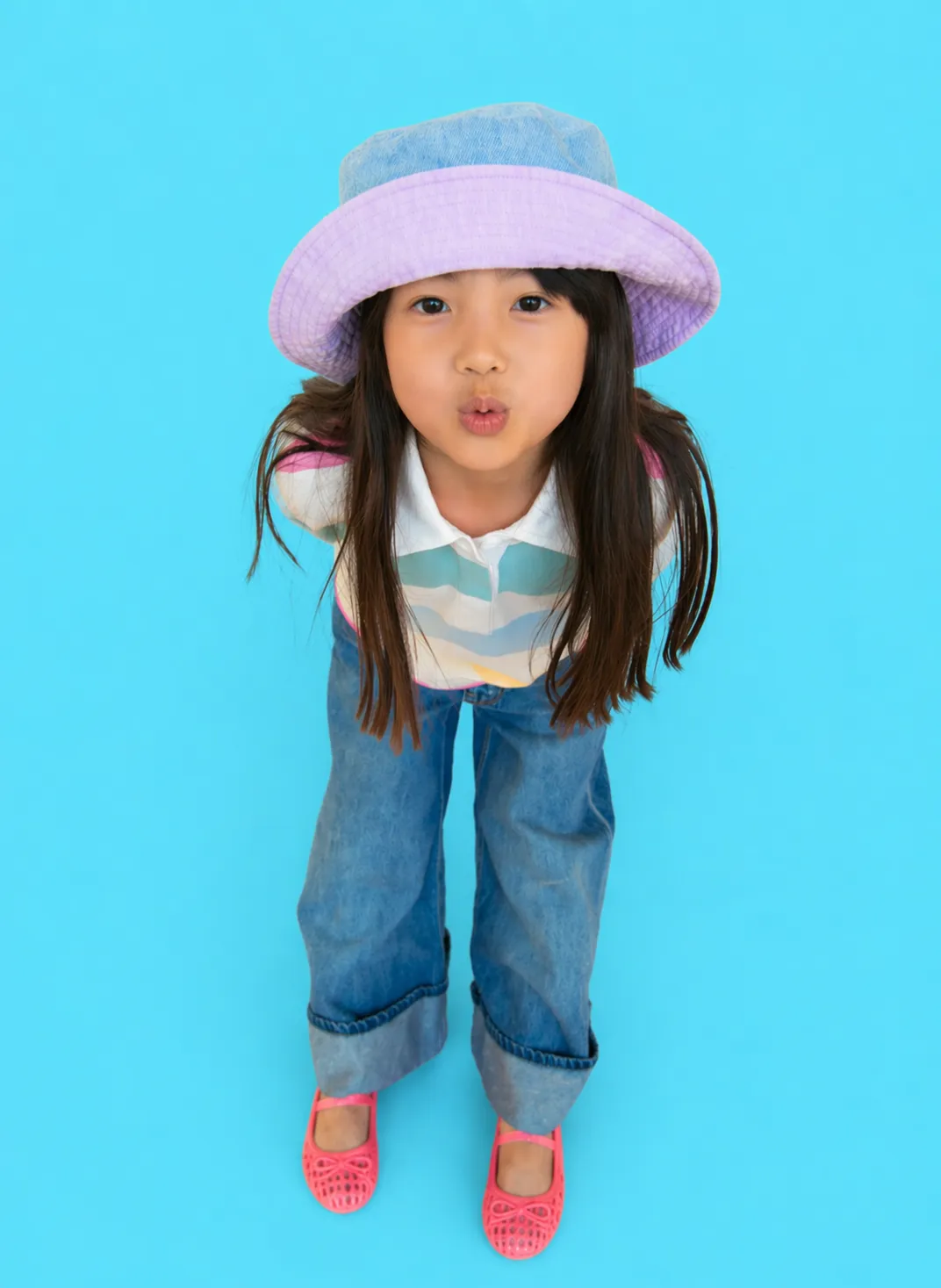Young girl with long dark hair wearing a purple bucket hat, striped shirt, wide-leg jeans, and pink shoes standing against a blue background and making a puckered facial expression.