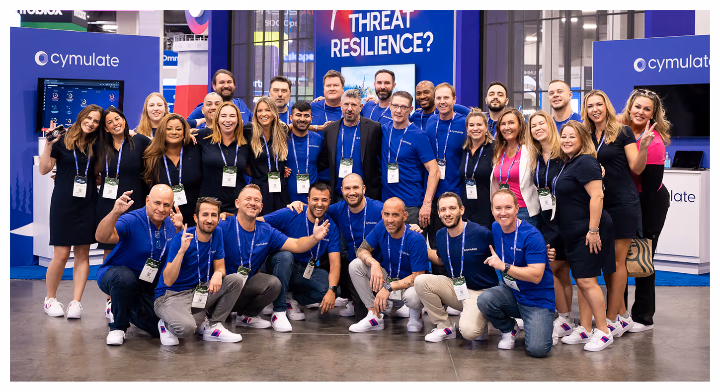Group of diverse colleagues posing together at a Cymulate booth with 'THREAT RESILIENCE?' sign in background.