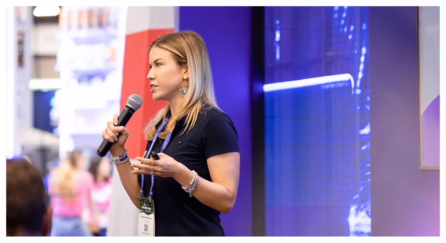Woman with blonde hair speaking into a microphone at a conference, wearing a black shirt and a badge.