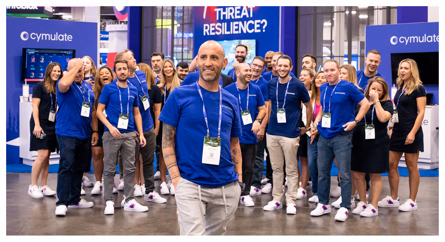 Group of people wearing blue Cymulate shirts and white sneakers standing and smiling at a conference booth with 'THREAT RESILIENCE?' sign.