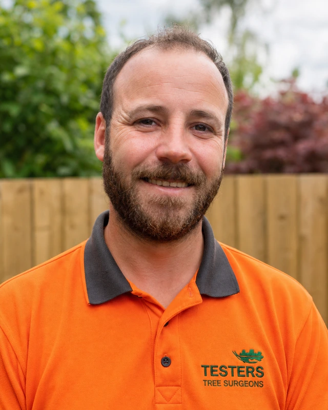 Smiling man with short hair and beard wearing an orange polo shirt with TESTERS TREE SURGEONS logo, standing outdoors with greenery and wooden fence in background.