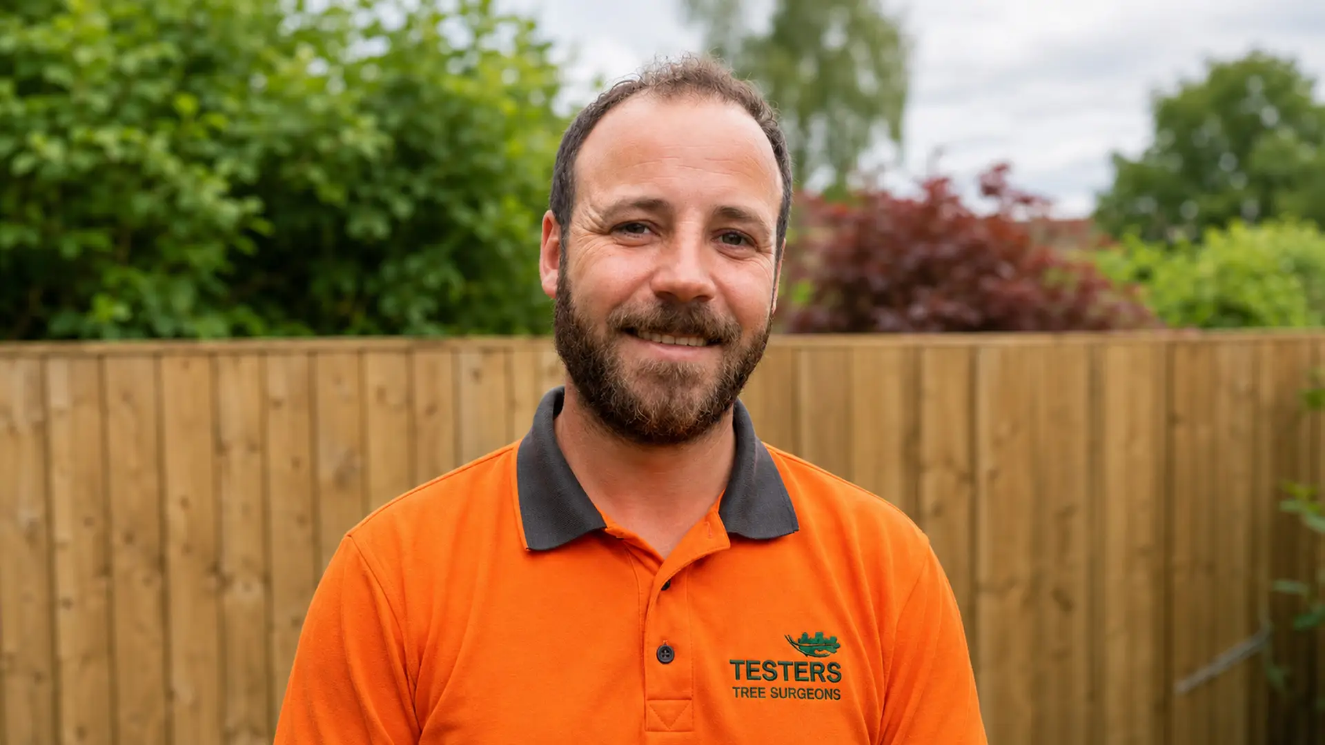 Smiling man with beard in orange shirt labeled Testers Tree Surgeons standing outside in backyard.
