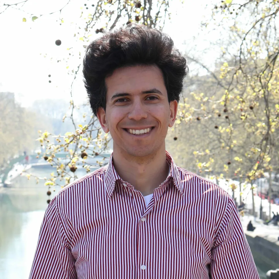 Smiling man with dark curly hair wearing a red and white striped shirt standing outdoors near a river and trees with sparse yellow leaves.