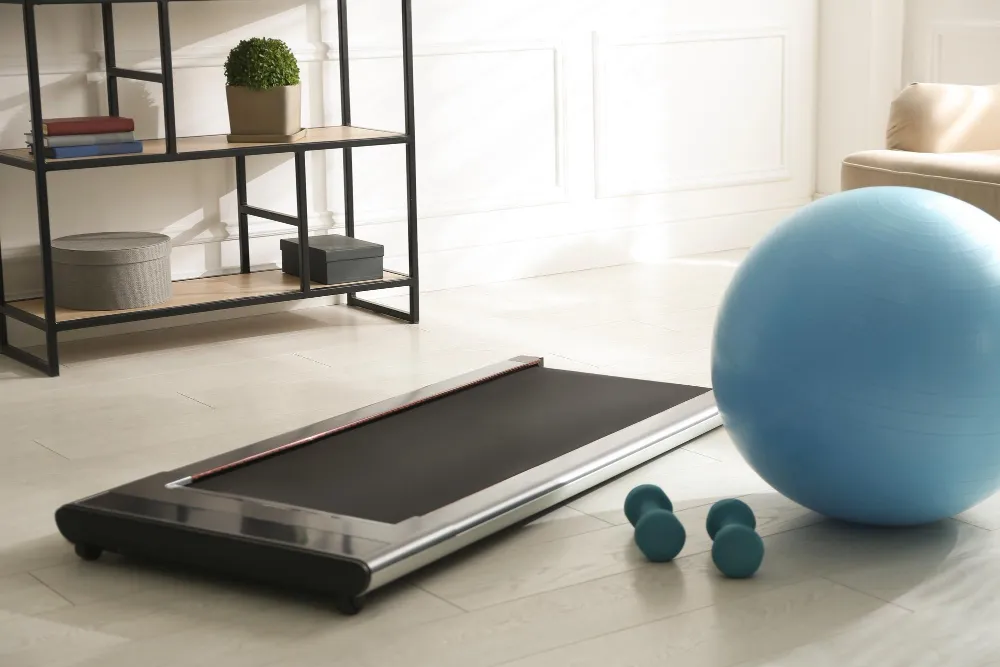 A flat walking pad treadmill with a blue exercise ball and small dumbbells in a bright, modern living space.