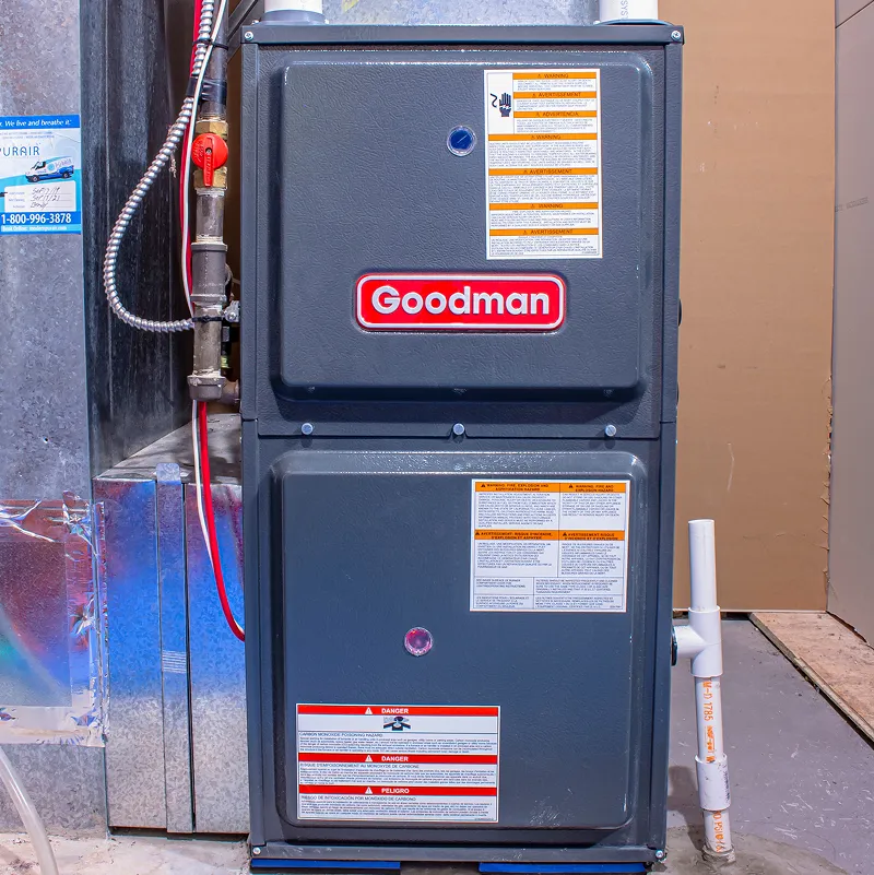 Close-up of a Goodman gas furnace with safety labels, installed in a basement utility area.