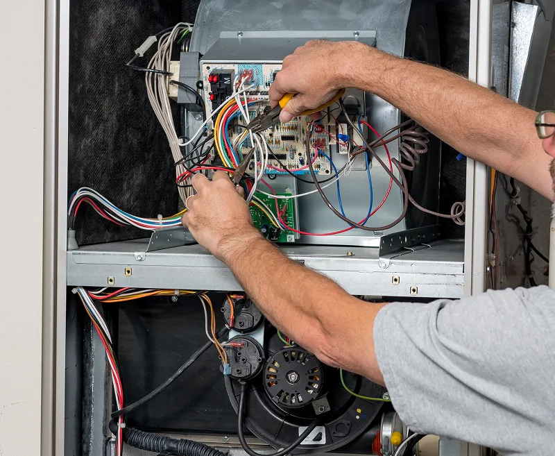 A technician using pliers to work on the wiring and control board inside an open furnace.