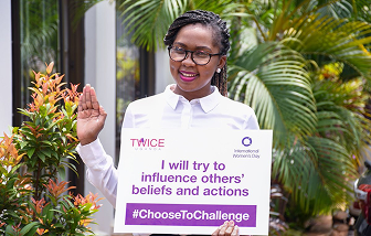 Woman in glasses and white shirt holding a sign that reads 'TWICE I will try to influence others' beliefs and actions #ChooseToChallenge' with International Women's Day logo.