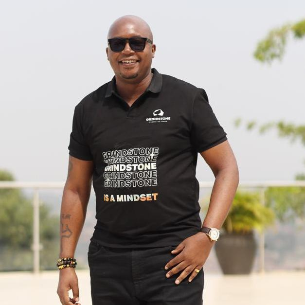 Smiling man wearing sunglasses and a black Grindstone polo shirt with the text 'Grindstone is a mindset', standing outdoors with a blurred background.