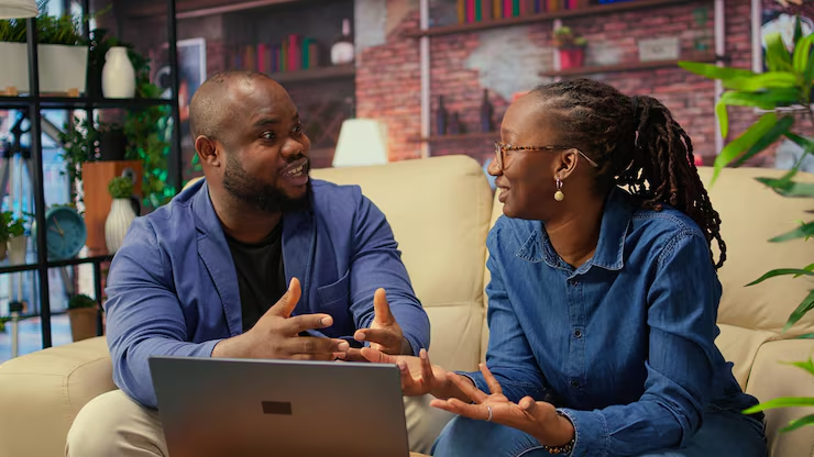 Two people engaged in a friendly discussion on a beige couch with a laptop in front of them in a cozy, plant-filled room.