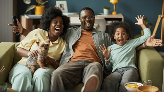 Happy family of three sitting on a green couch with a small dog, smiling and raising hands in excitement.