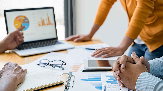 People in a business meeting reviewing charts, graphs, and a laptop with data visualizations on a wooden table.