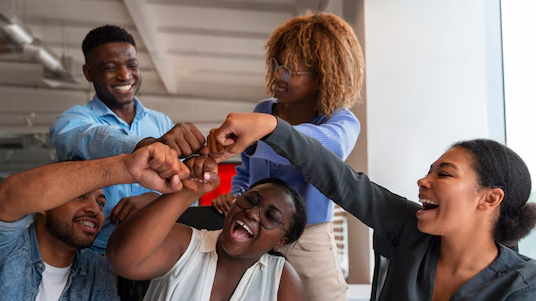 Group of five diverse young adults smiling and bumping fists in a celebratory gesture indoors.