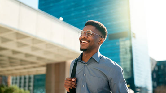 Smiling man wearing glasses and a blue shirt walking outdoors with a backpack in an urban setting.