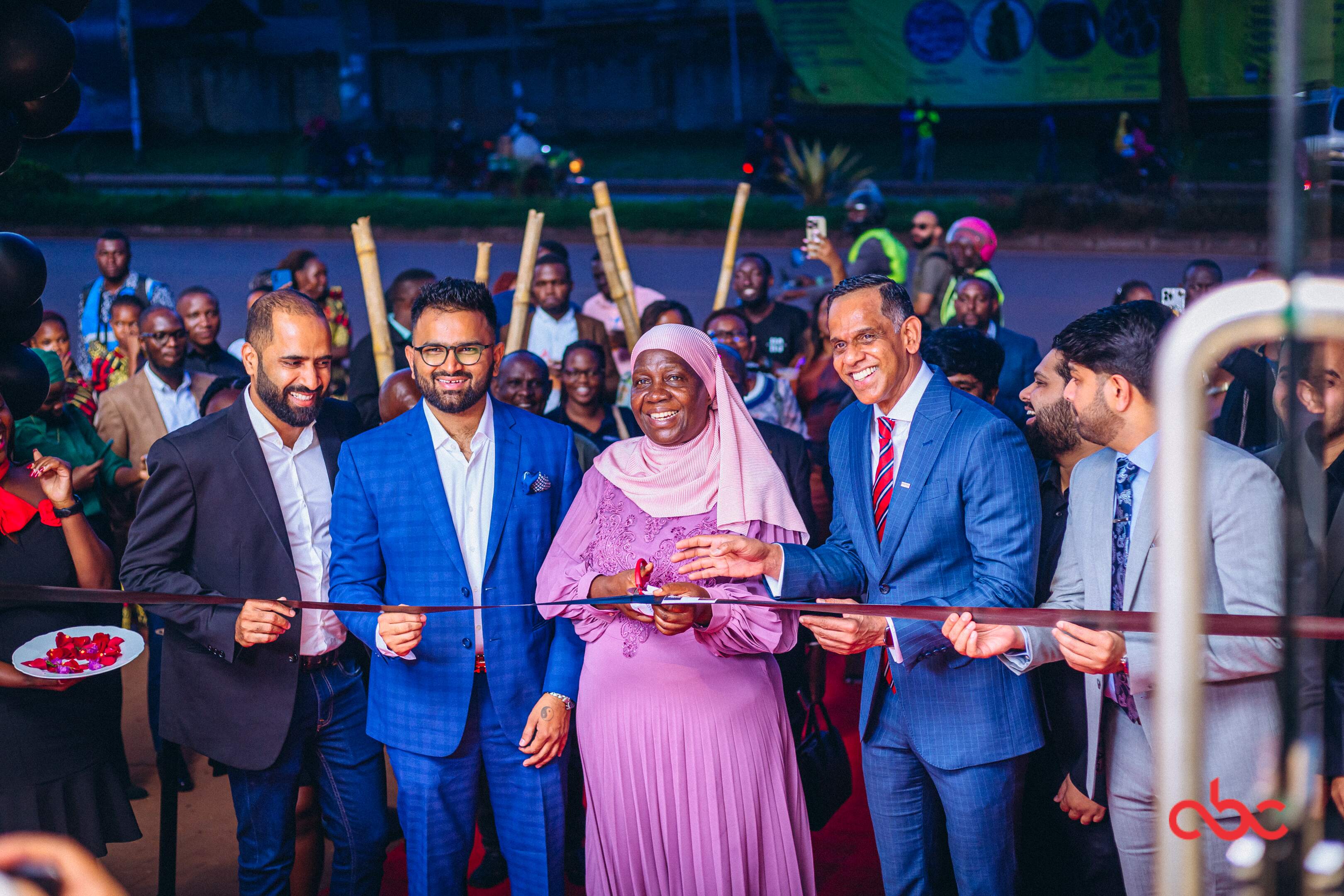 Group of people smiling and cutting a ribbon at an evening event with a crowd behind them.