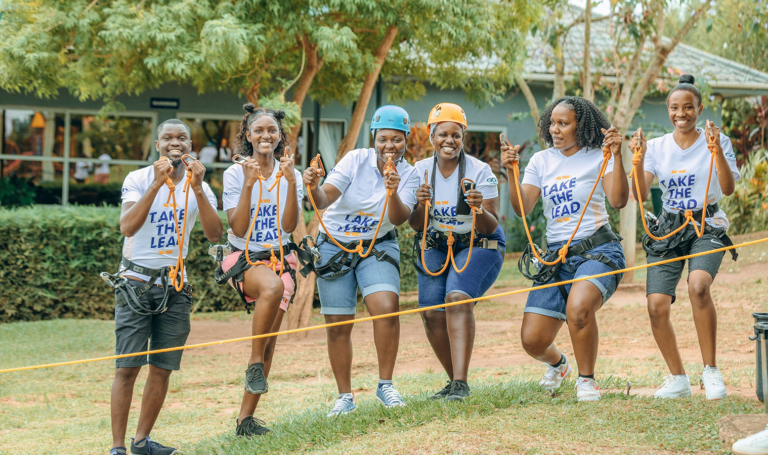 Six people in harnesses and helmets holding climbing ropes, smiling and posing outdoors during a team-building activity.