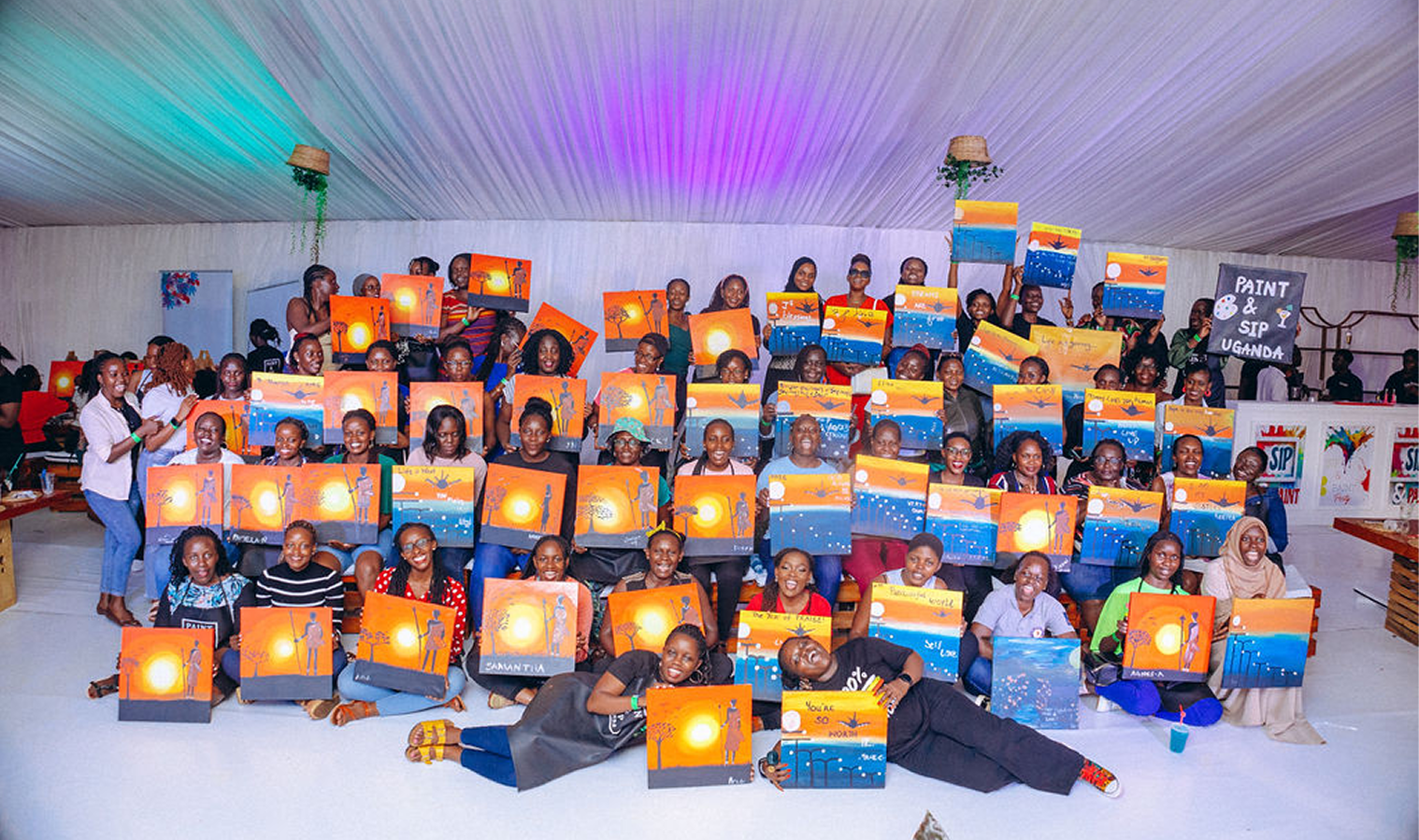 Group of diverse people posing indoors holding colorful sunset-themed paintings from a Paint & Sip Uganda event.