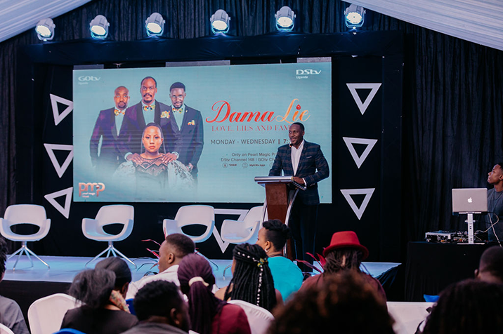 Man speaking at a podium in front of an audience with a large screen behind showing promotion for the TV show Dama Lie.