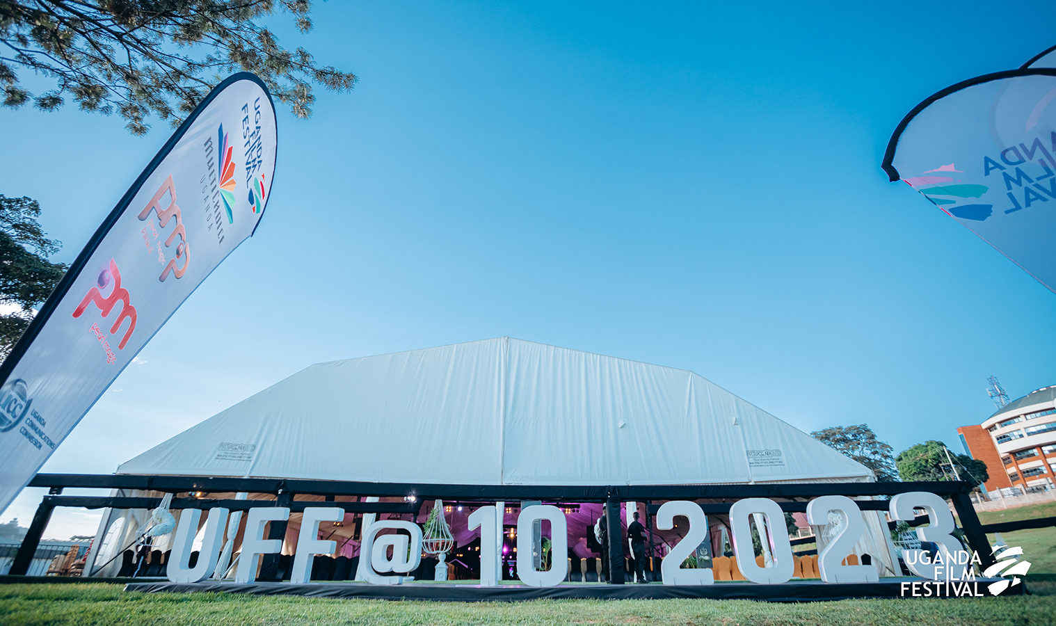 Large white tent with big letters spelling UFF @ 10 2023 on the grass in front, flanked by Uganda Film Festival flags.