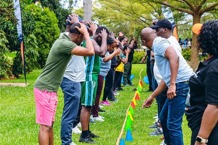 Group of people standing in two rows outdoors, facing each other with hands on their heads, participating in a team-building activity near colorful cones on the grass.