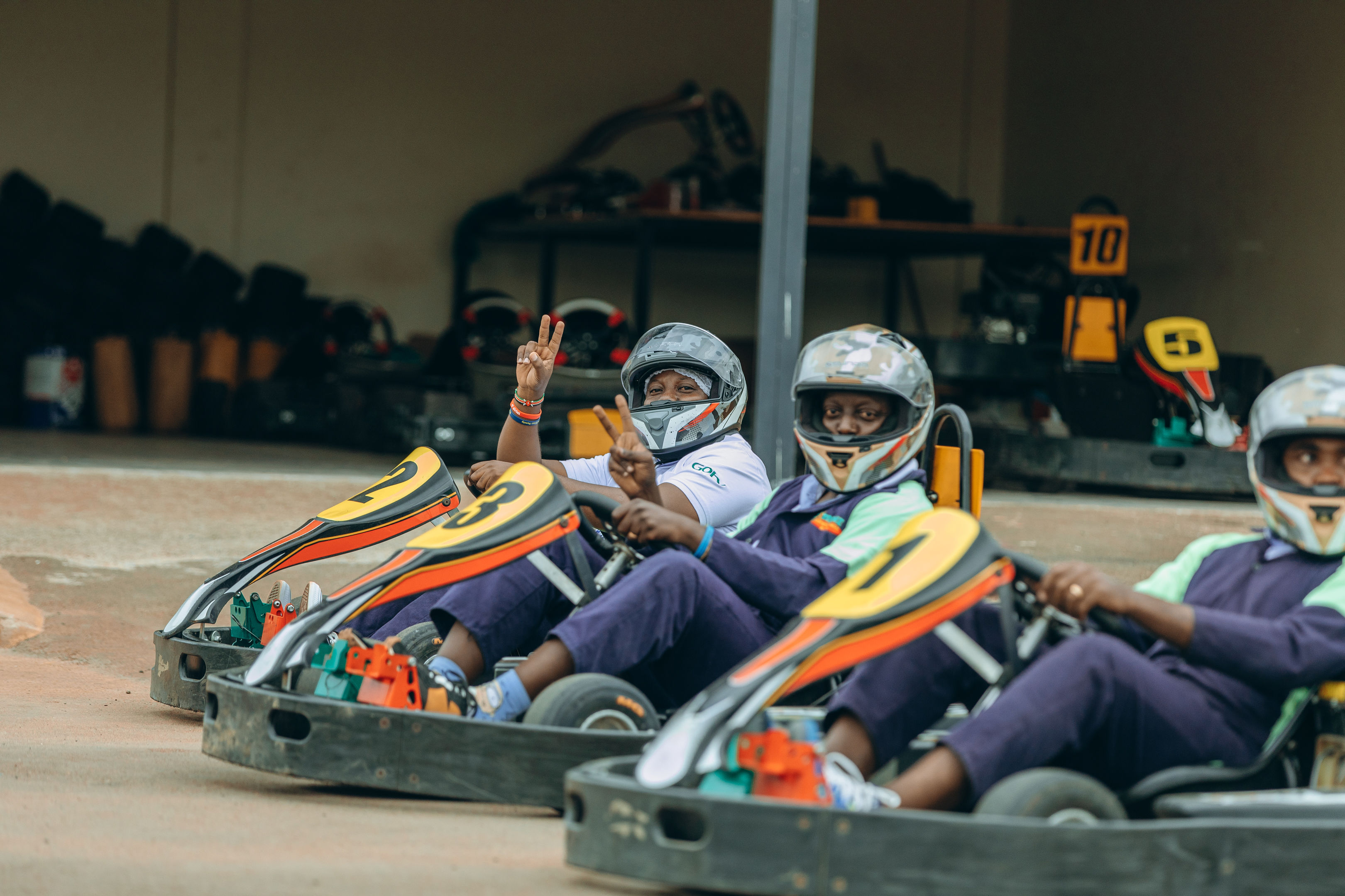 Three people wearing helmets seated in go-karts numbered 2, 3, and 1, two showing peace signs.