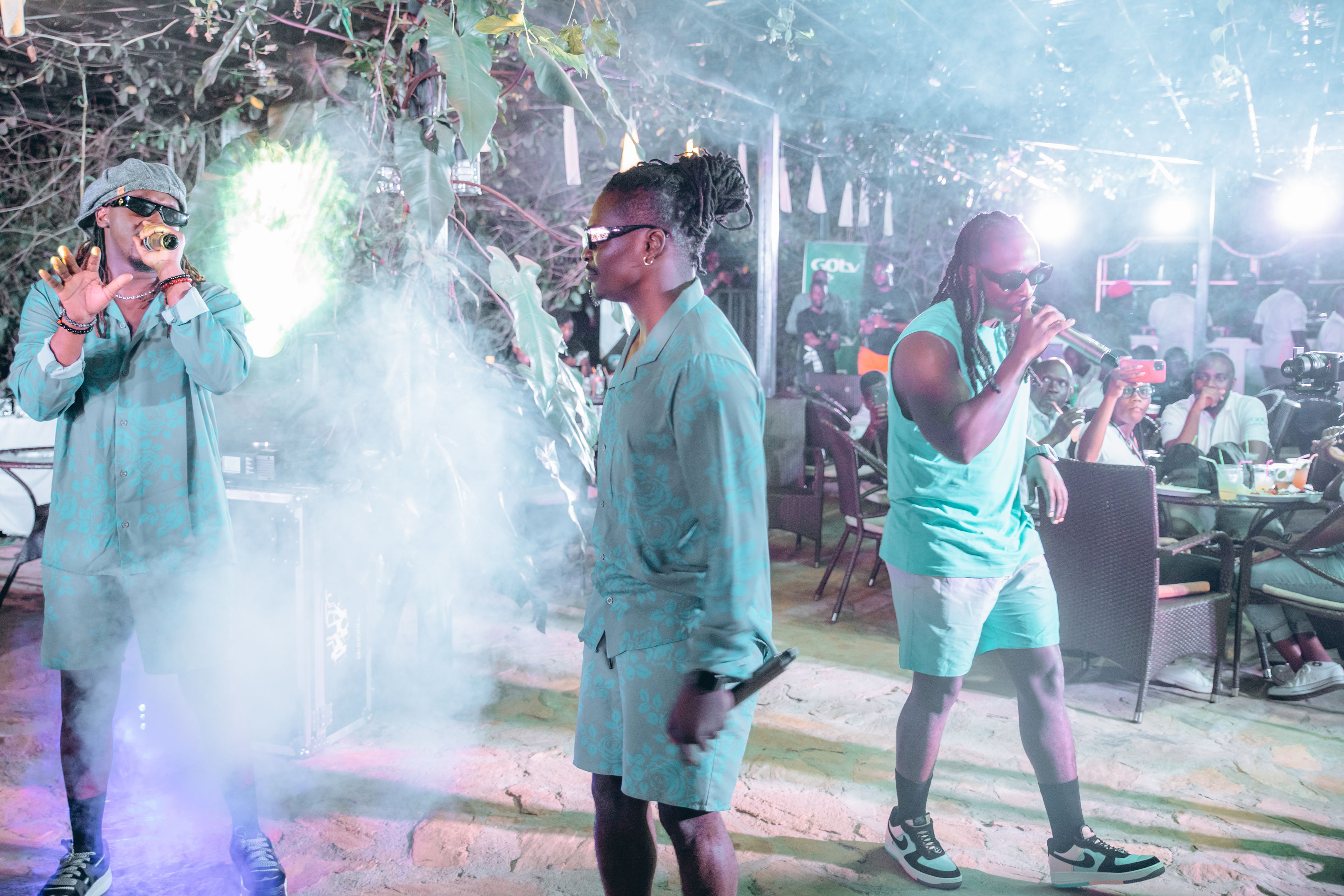 Three male performers with dreadlocks and sunglasses performing at an outdoor event with fog and seated audience in the background.