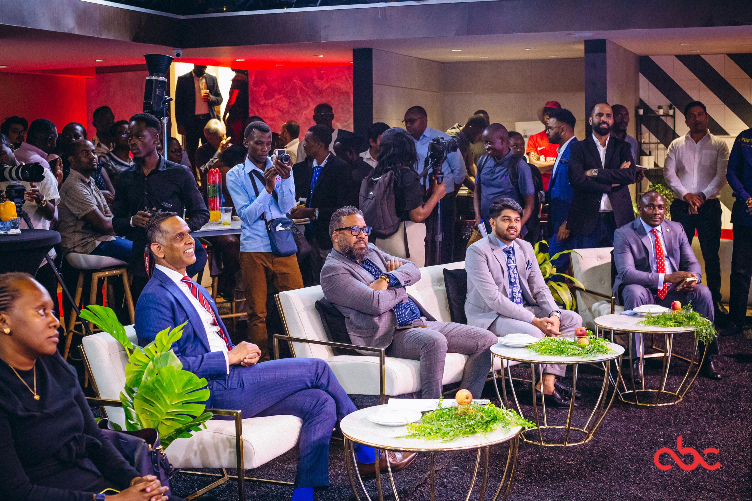 Group of well-dressed men sitting on white chairs with small tables decorated with greenery and apples, surrounded by an audience and photographers in an indoor event.