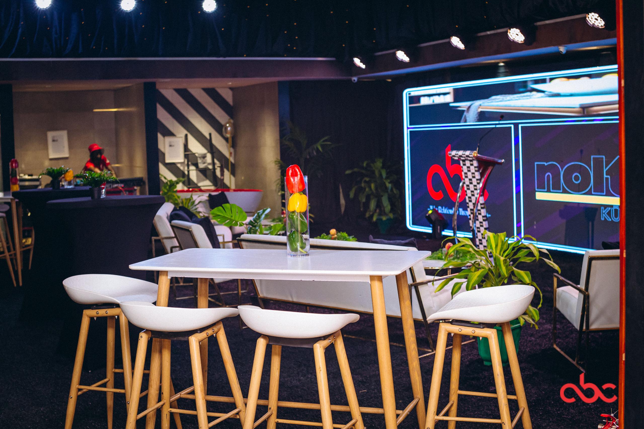 Modern event space with a white table and four wooden-legged stools, a clear vase with red and yellow tulips on the table, and a stage with a lectern and illuminated screens in the background.