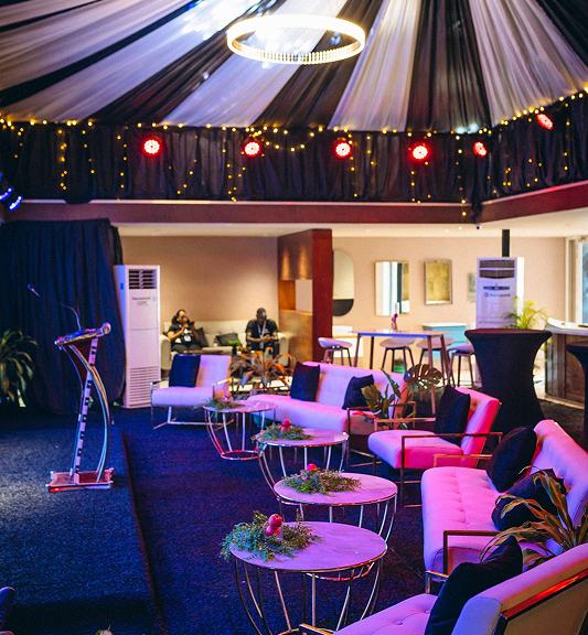 Interior event space with modern white sofas, circular tables decorated with plants, a microphone stand on a small stage, and warm overhead lighting under a draped ceiling.