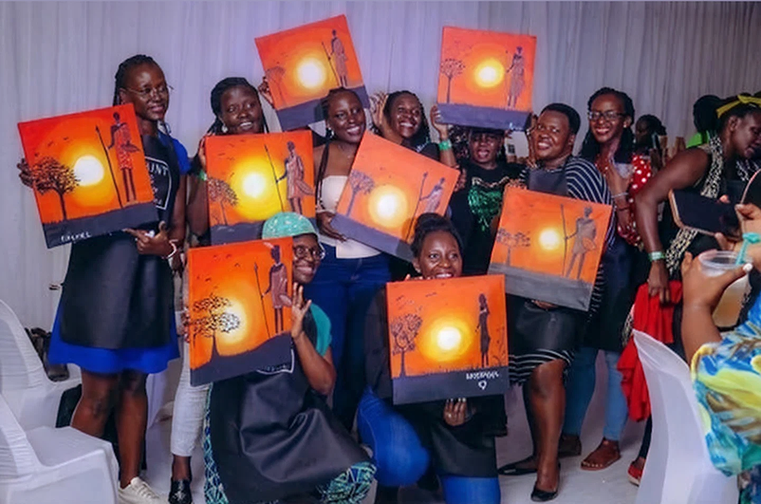 Group of people smiling and holding up their orange-themed paintings of a sunset with silhouetted figure and tree in a painting class setting.
