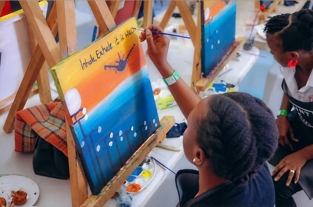 Two people engaged in painting on canvases set on wooden easels, one canvas showing a sunset with the words 'Inhale, Exhale, it is worth it.'