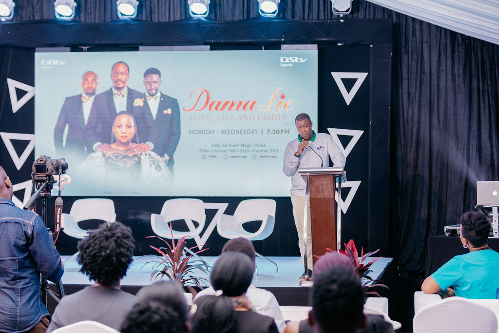 Man speaking at a podium during a presentation with an audience seated and a screen behind him showing promotional content for the TV show Dama Lie.