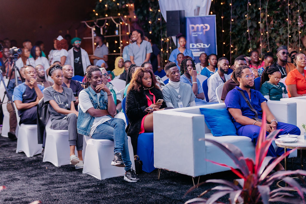 Audience seated indoors, attentively watching an event with some people sitting on white chairs and others on white sofas with blue cushions.