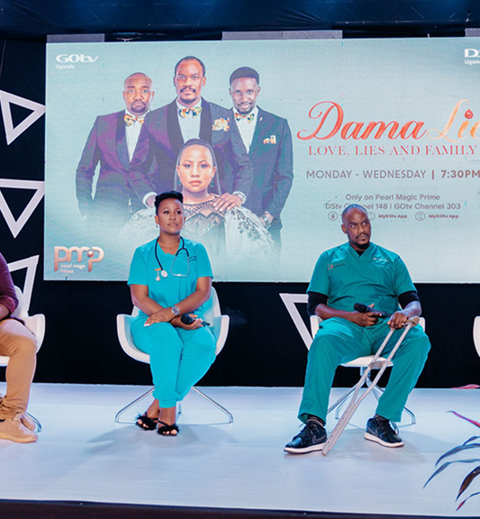 Two people in medical scrubs sitting on chairs in front of a large screen displaying a promotional poster for the show 'Dama Lie' with cast members dressed formally.