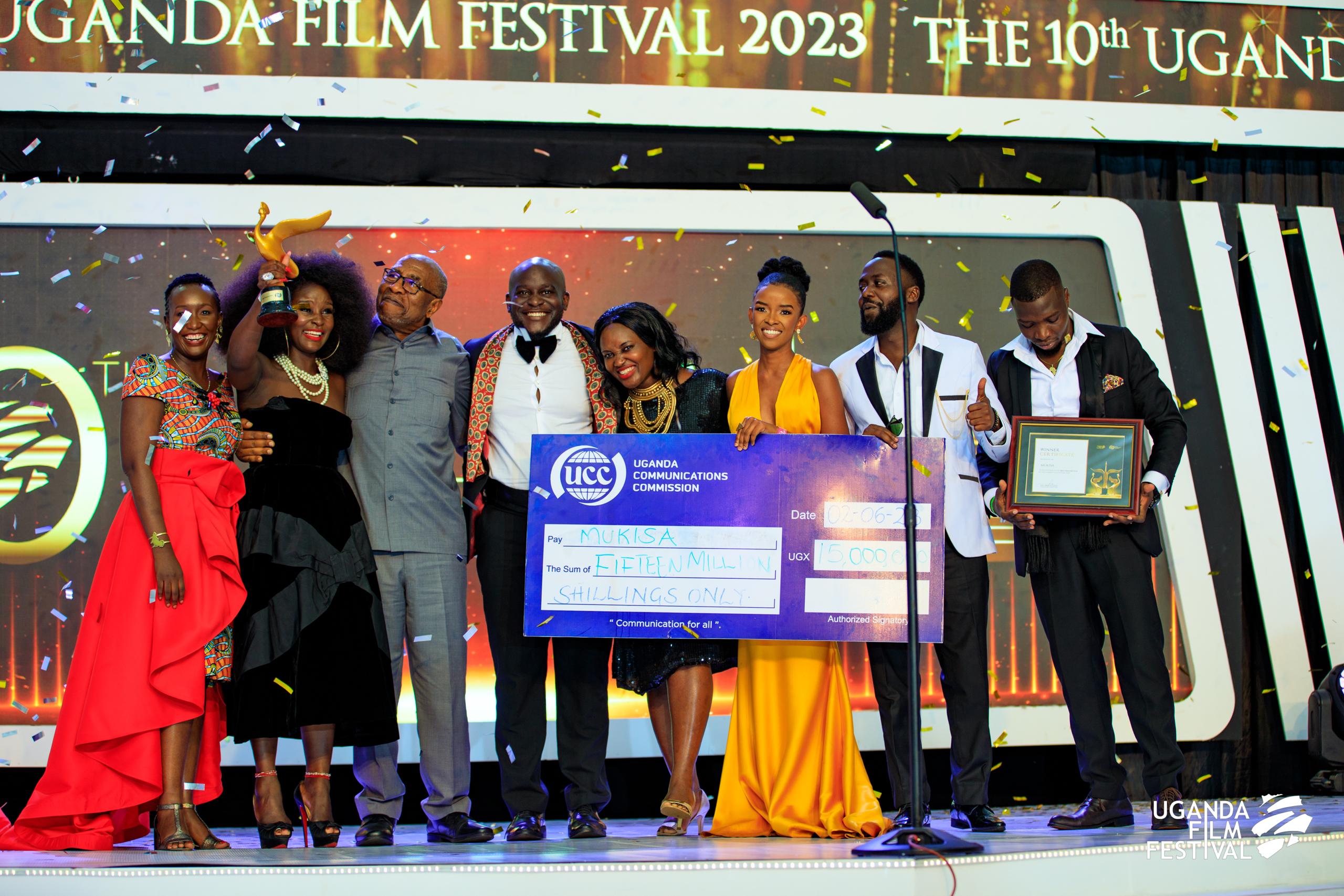 Group of eight people on stage at Uganda Film Festival 2023 celebrating with a large ceremonial check, trophy, and certificate.