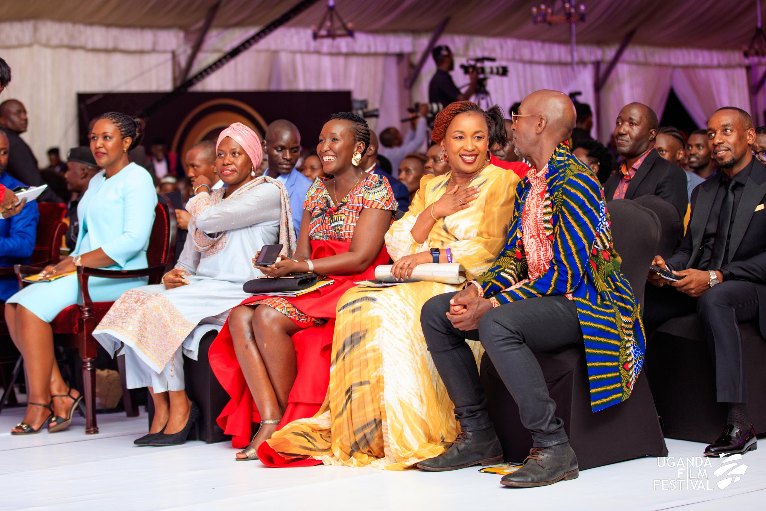 A group of well-dressed people seated indoors at the Uganda Film Festival, engaged in conversation and smiling.