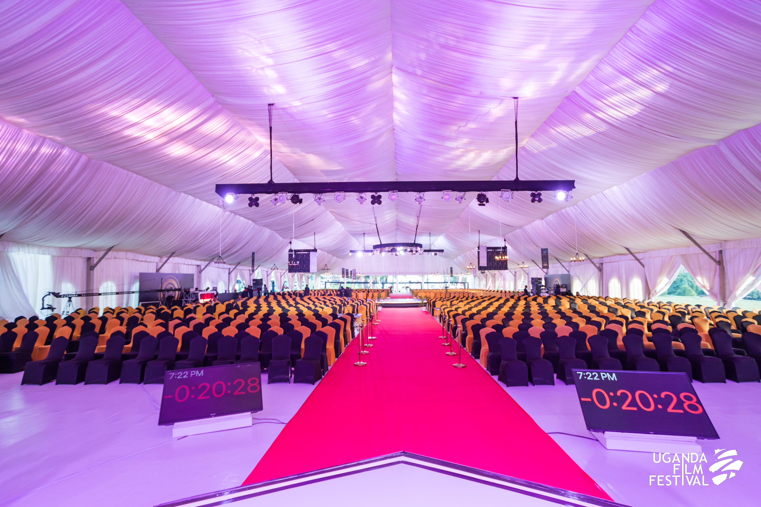 Large event tent with rows of black and orange covered chairs facing a stage, with a red carpet aisle and digital timers displaying countdown.
