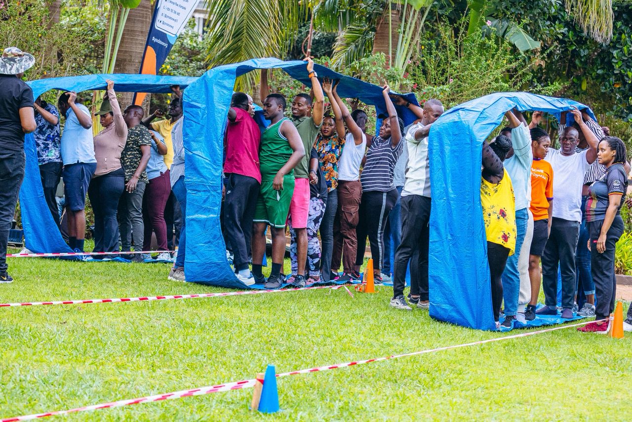 Group of people participating in an outdoor team-building activity holding up blue tarps while standing on grass.