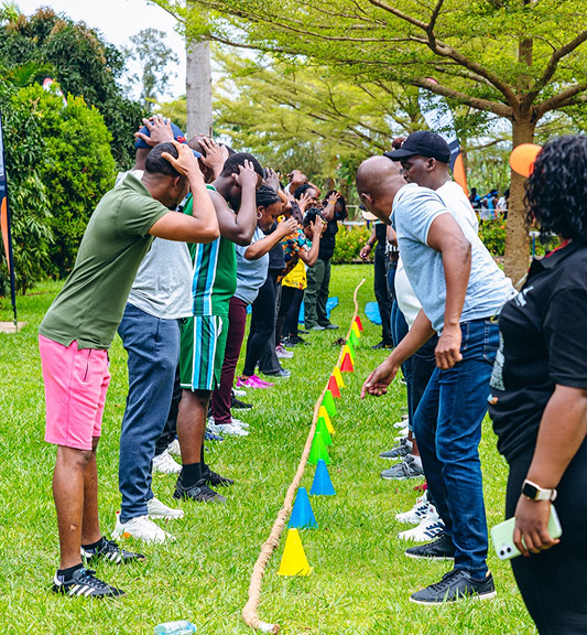 Two teams facing each other on grass with hands on heads, competing in a team-building outdoor activity marked by colorful cones and a rope.