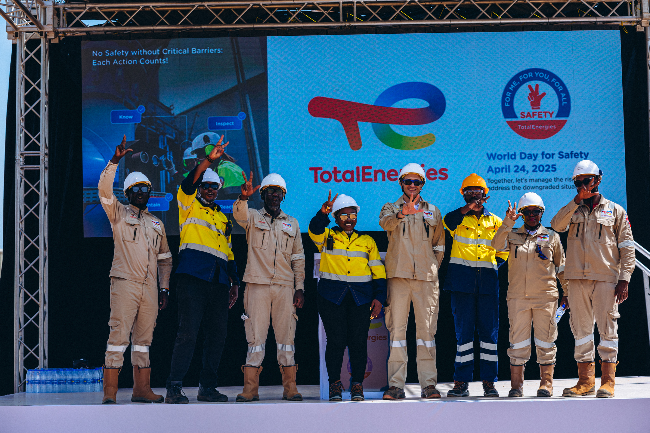 Eight TotalEnergies workers wearing safety gear stand on stage showing peace signs in front of a World Day for Safety 2025 banner.